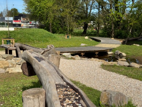 Speelplaats met vlonderpad, waterplas en een houtenspeeltoestel met water in een groene omgeving met bomen.