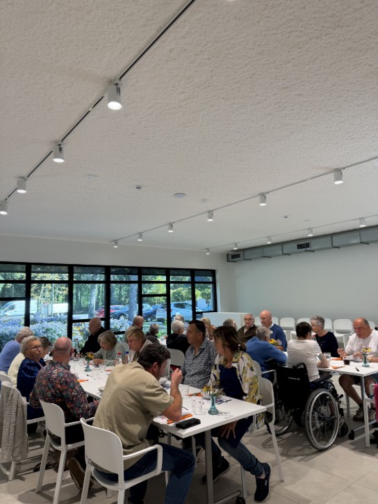 Een groep ouderen zit samen aan tafels in een lichte, moderne eetruimte met grote ramen. (vergroot foto)