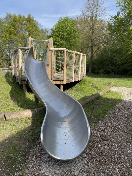 Gladde metalen glijbaan met houten hekwerk in een groene speeltuin bij zonnig weer.