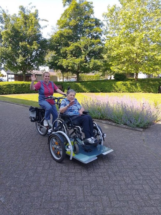 Een vrouw fietst en vervoert een oudere dame in een rolstoel op een speciale fiets, beiden zwaaien vrolijk.