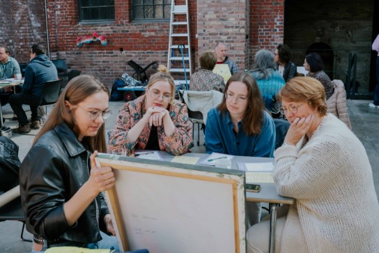 Vier vrouwen zitten buiten rond een tafel en bekijken samen een schilderij of kunstwerk. (vergroot foto)