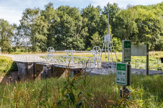 Fietsroutebordjes bij een kleine metalen brug in een groene, bosrijke omgeving. (vergroot foto)