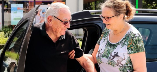 Een vrouw helpt een oudere man uit een auto, beiden glimlachen. (vergroot foto)