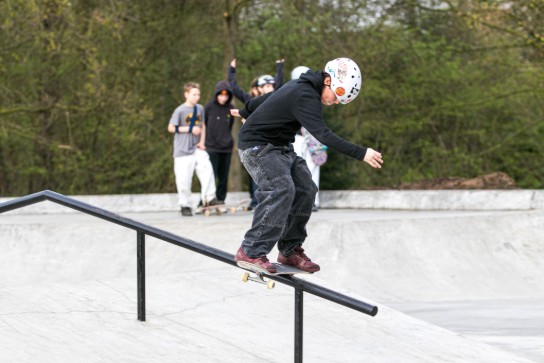 Jongere met helm skate op een rail in een skatepark, terwijl anderen toekijken.