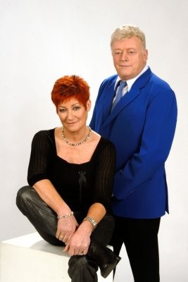 Portret van een vrouw met rood haar zittend en een man in een blauw jasje staand, beide kijken naar de camera. (vergroot foto)