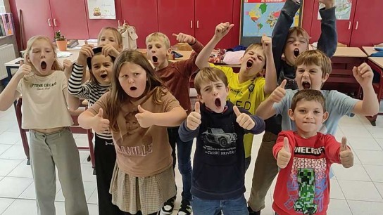 Een groep enthousiaste kinderen in een klaslokaal steekt duimen omhoog en juicht. (vergroot foto)